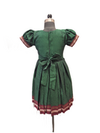 A green patterned dress displayed on a dress form mannequin against a white background. The dress features short puff sleeves with red and white patterned cuffs, a matching waist tie, and a pleated skirt with a red and white patterned hem. - House of Kritee