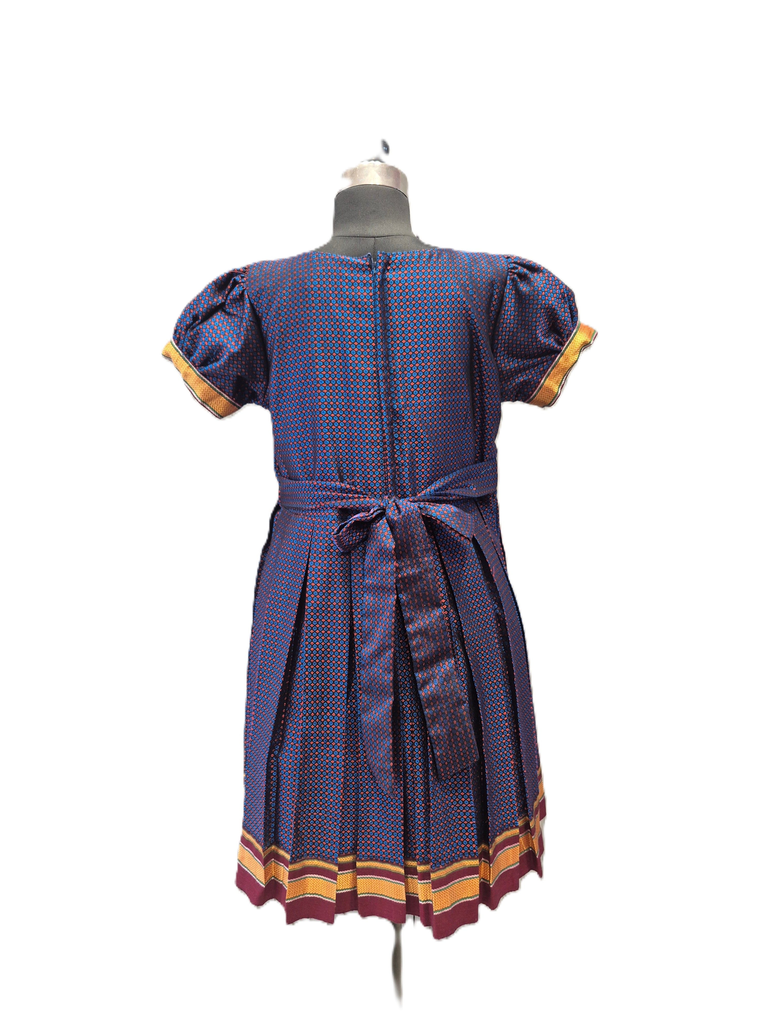 A short-sleeve dress displayed on a mannequin from the back, featuring a blue and red patterned fabric with yellow and burgundy striped cuffs and hem. - House of Kritee