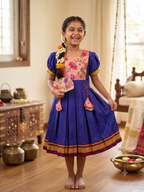 A young girl stands barefoot in a traditional Indian room wearing a royal blue cotton saree-style dress with a pink floral embroidered bodice, yellow sleeve accents, and gold zari borders. She is holding a matching pink and gold hand fan. - House of Kritee