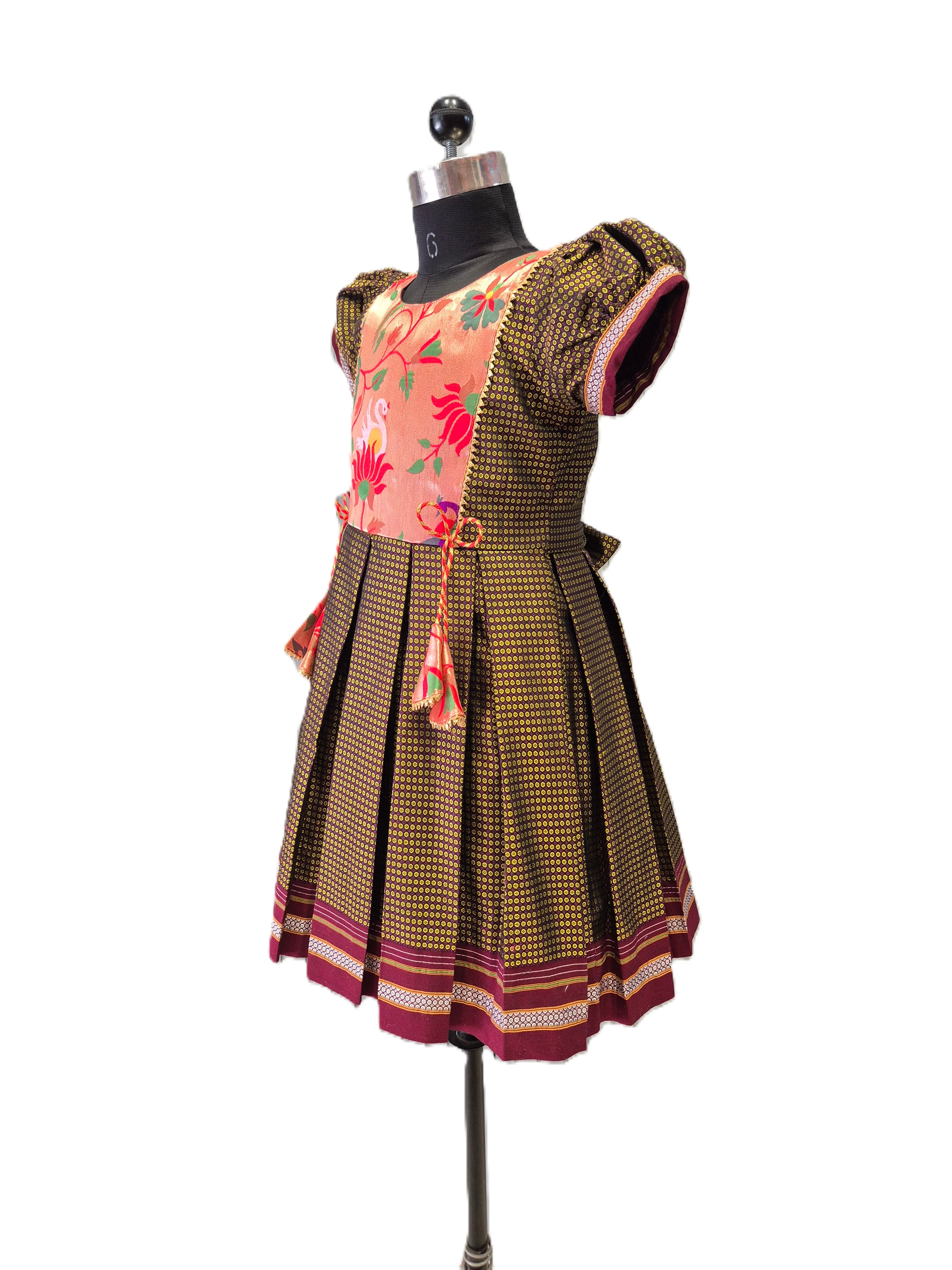 A traditional Indian salwar suit set displayed on a black dress form mannequin. The set features a brown and gold patterned pleated skirt with a maroon border, a matching patterned kurta sleeve, and a peach-colored floral printed bodice with a tassel tie. - House of Kritee