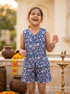 Happy Desi digital girl smiling in a premium blue and pink floral print cotton waistcoat and shorts co-ord set against a blurred warm cream garden backdrop.