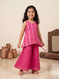 A young girl smiling and posing in a bright pink traditional Indian outfit with vertical metallic gold stripes, featuring a sleeveless tunic top and wide-leg pants, set against a warm beige background with decorative copper pots and a mirror. - House of Kritee