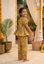 A young girl smiling and looking back over her shoulder while wearing a matching yellow and green printed two-piece outfit with a peplum top and wide-leg pants, standing in an outdoor courtyard decorated with garlands and potted plants.
