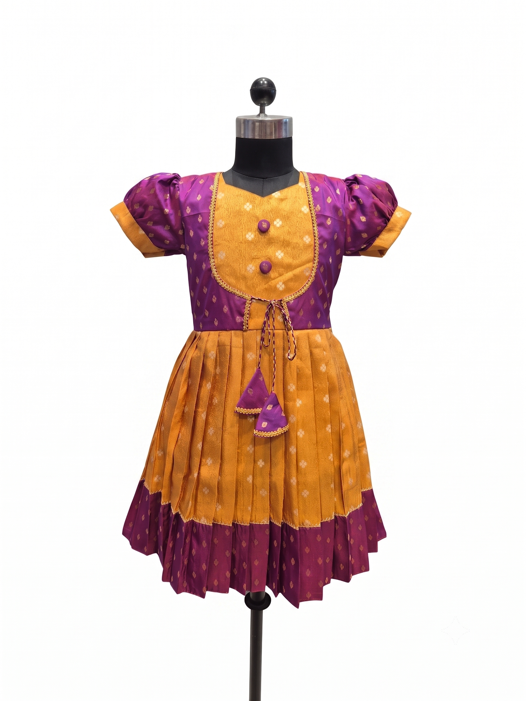 A traditional Indian girl's dress displayed on a black dress form mannequin. The dress features a purple bodice with gold polka dots, short puff sleeves, a gold embroidered chest panel with three purple buttons, and a pleated skirt in alternating yellow and purple patterned fabric - House of Kritee