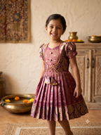 A young girl smiling and wearing a traditional Indian salwar kameez dress in a warm-toned indoor setting. The dress is a dusty rose/mauve color with gold zari embroidery and patterned details, featuring puff sleeves, a buttoned yoke, and a pleated skirt. - House of Kritee
