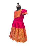 A traditional Indian girl's dress displayed on a black dress form mannequin. The dress features a bright magenta pink body with a contrasting orange and gold patterned yoke and a matching pleated skirt. - House of Kritee