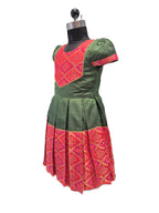 A traditional Indian girl's dress displayed on a black dress form mannequin. The dress is olive green with a bright pink and gold patterned yoke and a matching pleated skirt border. - House of Kritee