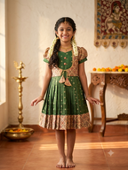 A young girl with braided hair stands barefoot indoors wearing a green and gold traditional dress with a pleated skirt and patterned border. The background shows a warm-toned room with a brass lamp, carved furniture, and wall decor. - House of Kritee