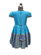 A blue short-sleeve dress shown from the back on a dress form mannequin, featuring a pleated skirt with a blue, gold, and green patterned border. - House of Kritee