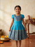A young girl smiling and wearing a traditional Indian blue and gold patterned dress, standing in a warm, decorated indoor setting with a wooden floor and decorative brass vessels. - House of Kritee
