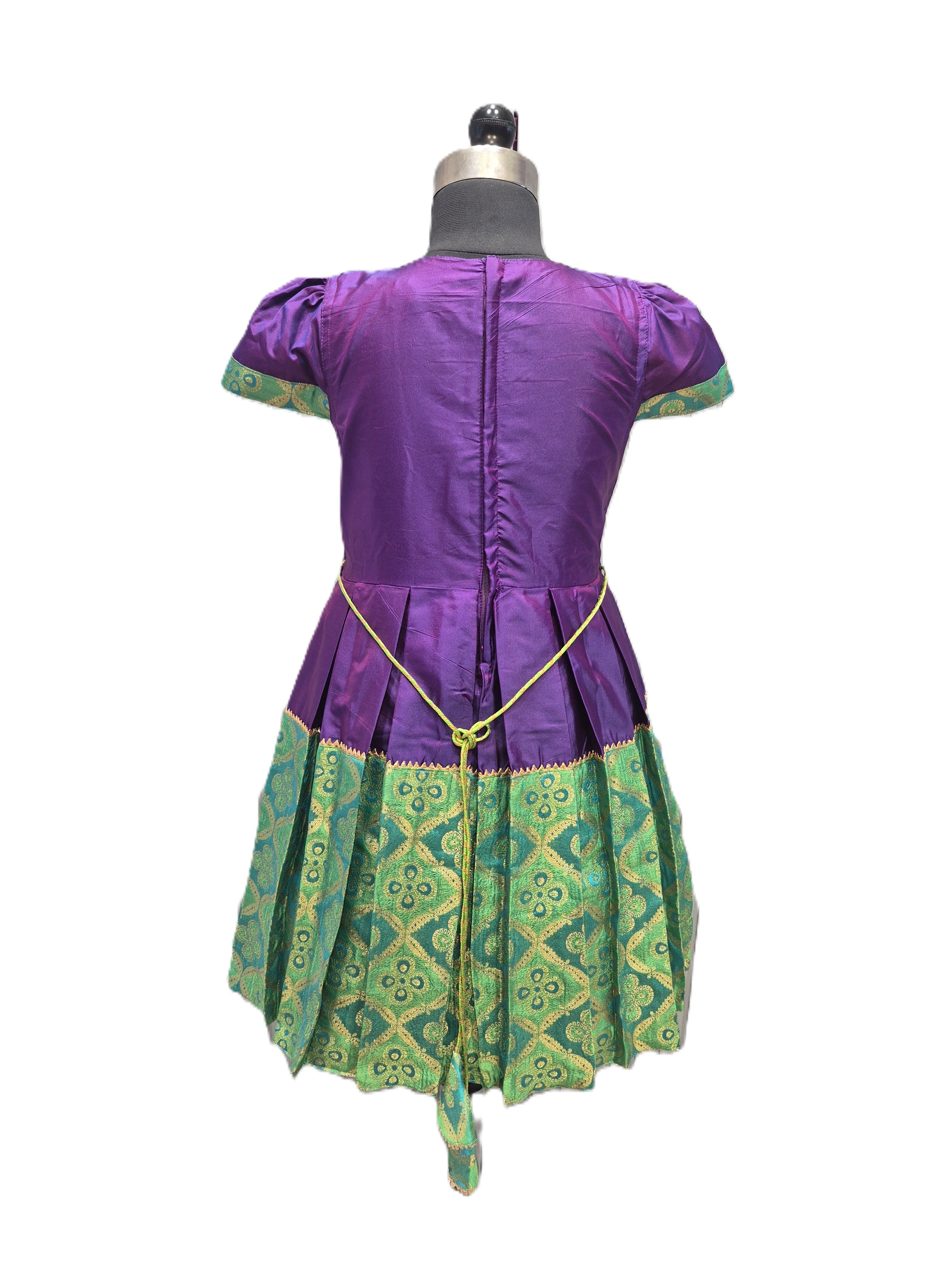 A purple and green traditional-style dress displayed on a dress form against a white background. The back view shows a purple bodice with short sleeves, a gold chain detail at the waist, and a green patterned skirt. - House of Kritee