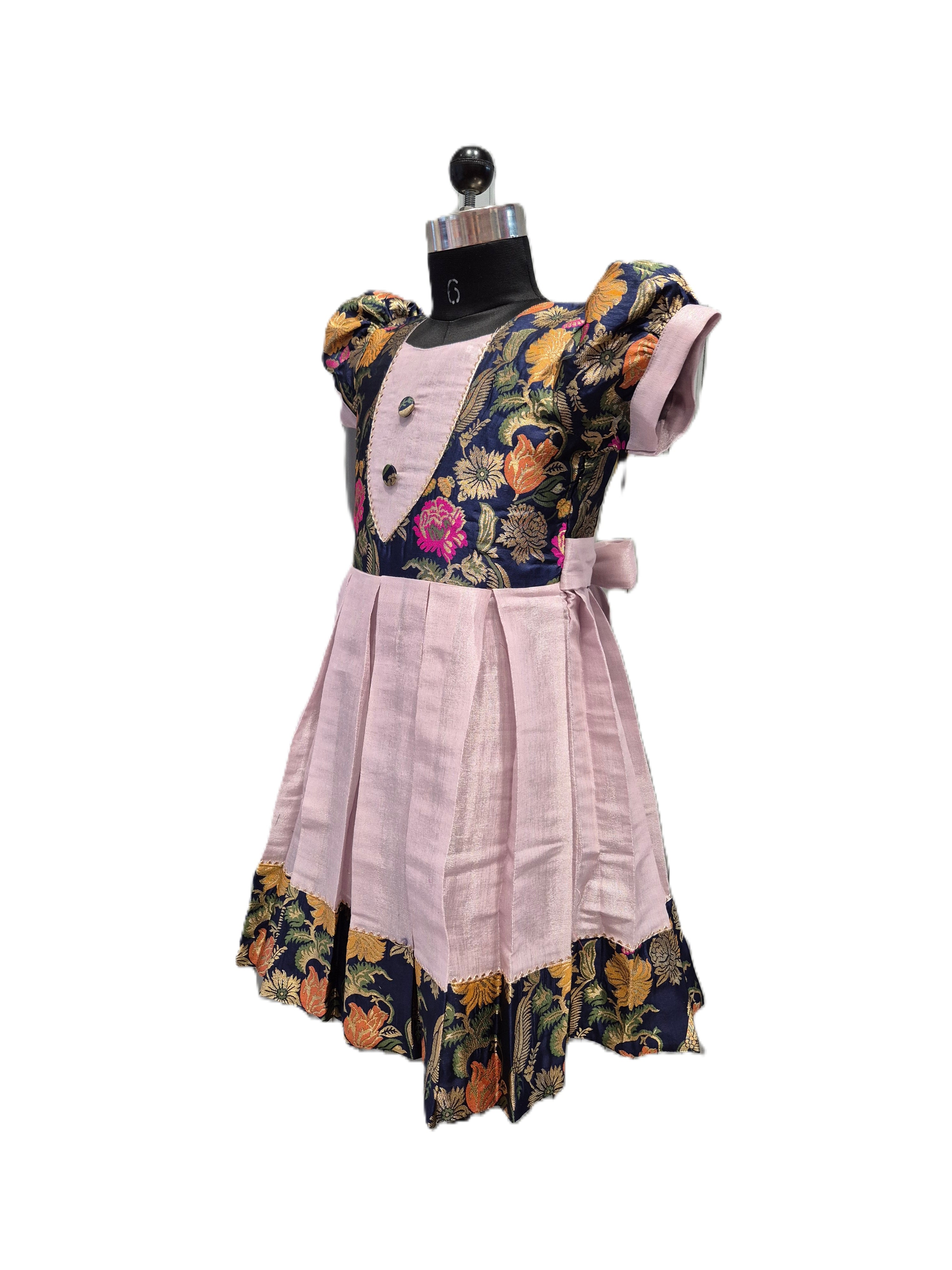 A vintage-style children's dress displayed on a black dress form mannequin against a white background. The dress features a light pink linen skirt and bodice, with a dark navy blue floral patterned yoke and ruffled trim. - House of Kritee