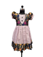 A children's dress displayed on a black dress form mannequin against a plain white background. The dress has a light pink pleated skirt, a navy blue bodice with pink floral embroidery, and gold patterned sleeves and hem. - House of Kritee