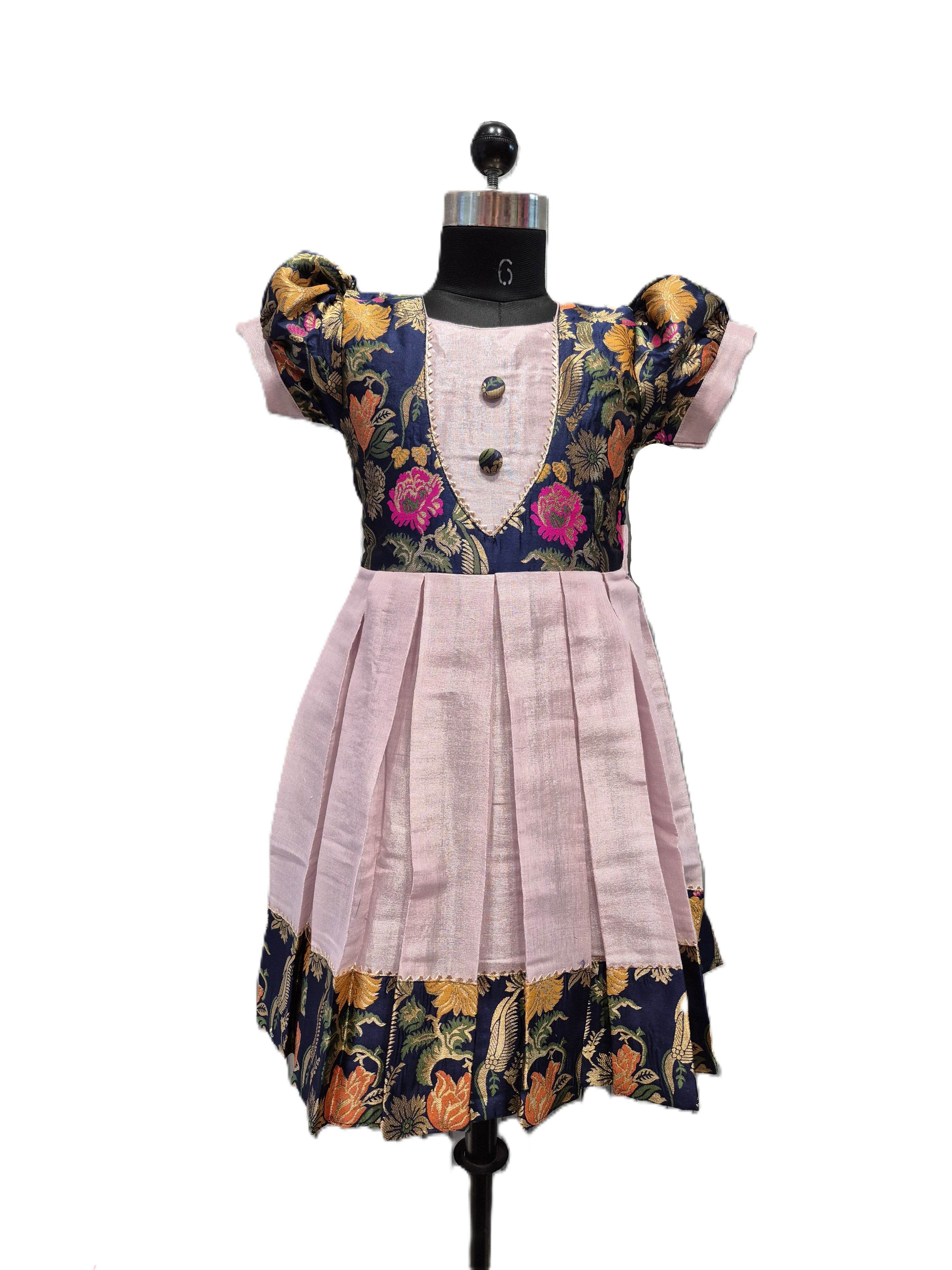 A children's dress displayed on a black dress form mannequin against a plain white background. The dress has a light pink pleated skirt, a navy blue bodice with pink floral embroidery, and gold patterned sleeves and hem. - House of Kritee