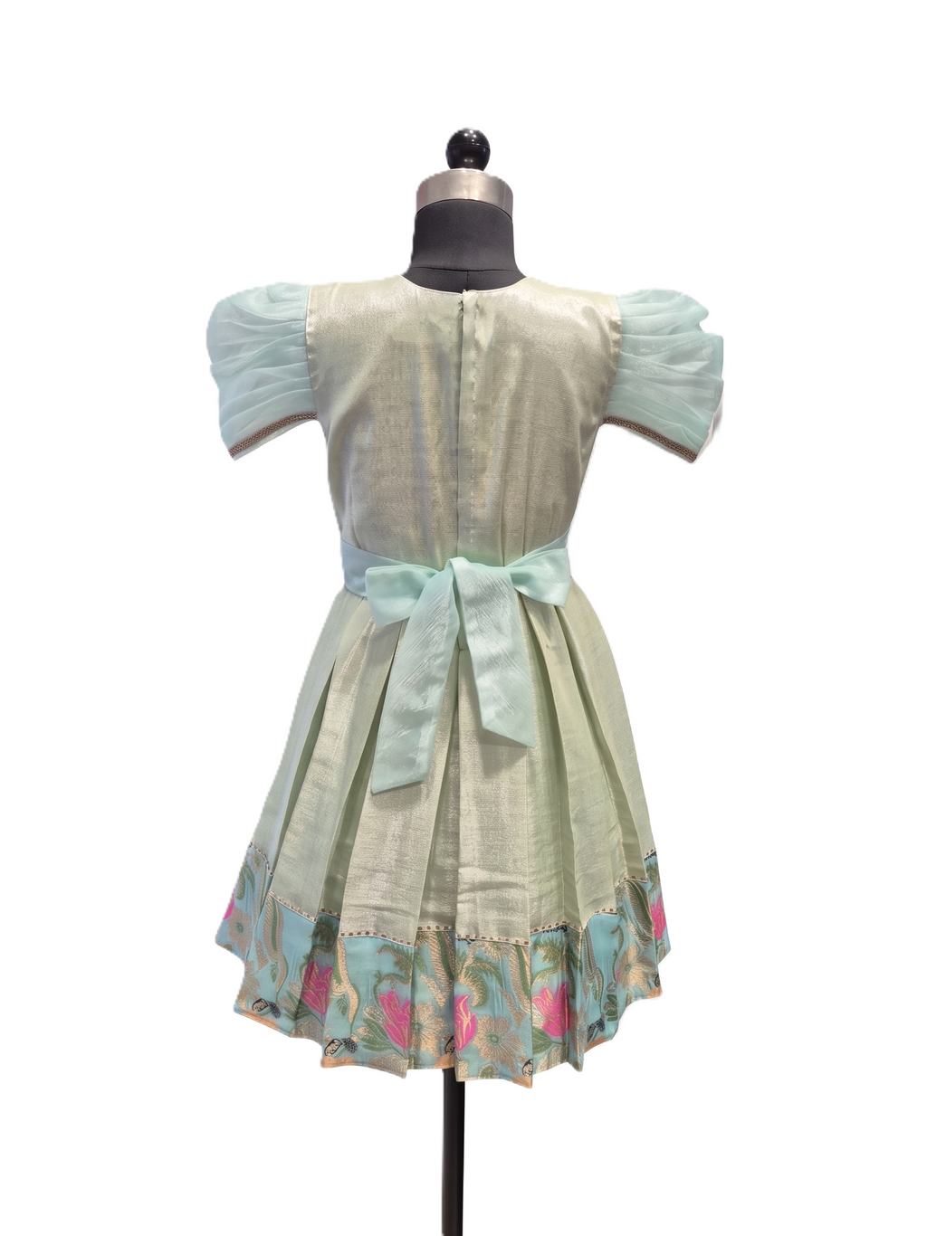 A light sage green dress displayed on a black dress form mannequin against a white background. The dress features short puff sleeves, a matching waist bow, and a floral patterned hem with pink and blue details. - House of Kritee