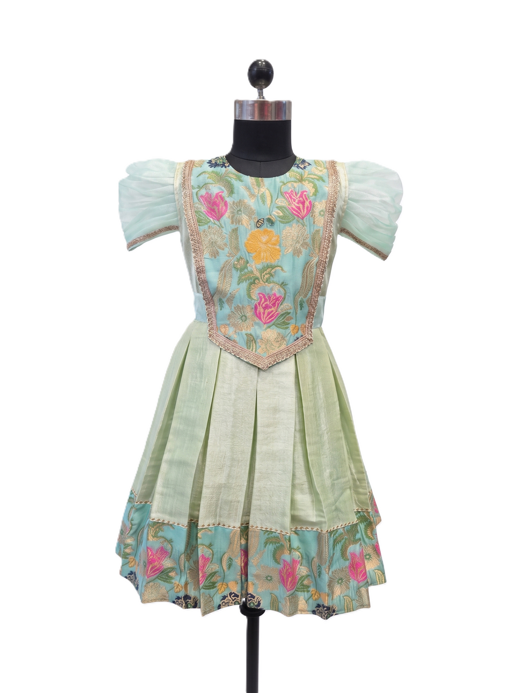 A light green and turquoise embroidered dress displayed on a black dress form mannequin against a white background. The dress features puff sleeves, a light green pleated skirt, and a turquoise bodice decorated with pink and yellow floral embroidery and gold trim. - House of Kritee