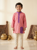 A young boy smiling and standing barefoot indoors, wearing a pink traditional kurta with a purple patterned collar and hem detail, paired with cream pants. - House of Kritee