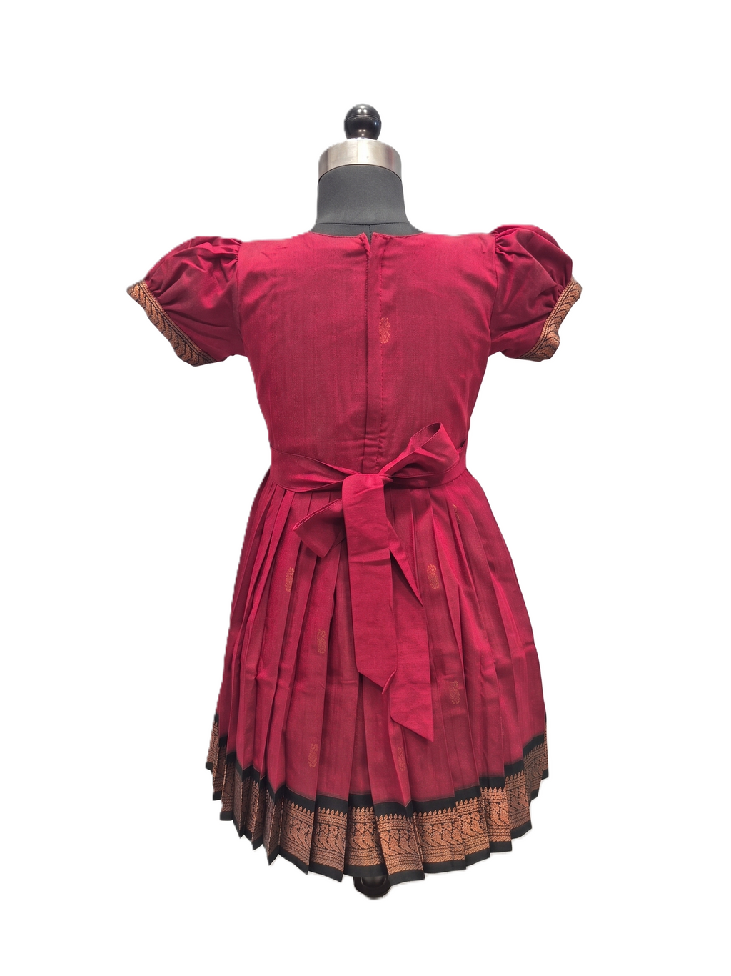 A red cotton dress with puff sleeves and a matching waist tie, displayed on a mannequin from the back. The dress features a pleated skirt and a decorative gold and black patterned border along the hem. - House of Kritee