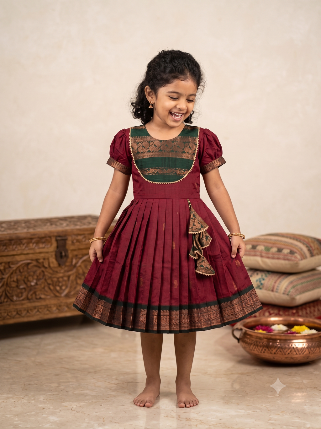 A young girl smiling and twirling in a traditional Indian salwar kameez dress. The dress is maroon with a dark green and gold embroidered yoke and a matching pleated skirt with a decorative ruffle. - House of Kritee