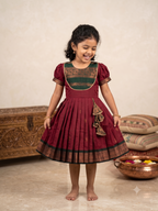 A young girl smiling and twirling in a traditional Indian salwar kameez dress. The dress is maroon with a dark green and gold embroidered yoke and a matching pleated skirt with a decorative ruffle. - House of Kritee