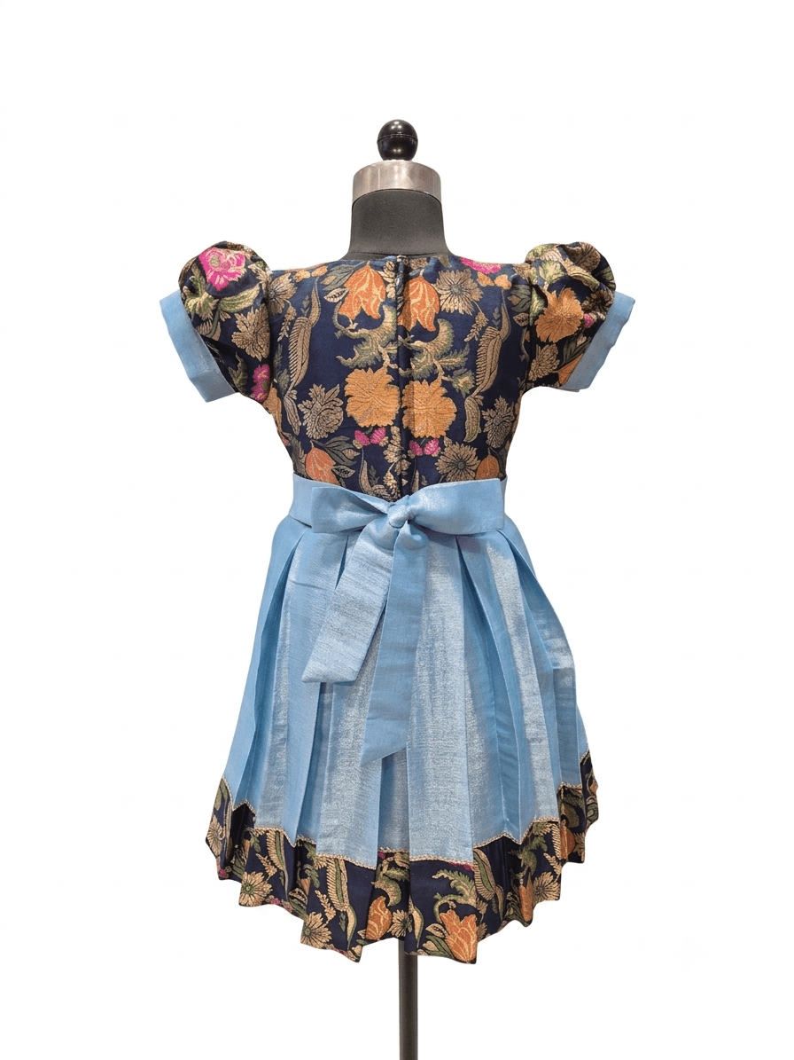 A short-sleeve dress displayed on a mannequin against a plain white background. The dress has a dark navy floral bodice with orange, yellow, and pink flowers, light blue puff sleeves, a matching light blue waist tie, and a light blue flared skirt with a dark floral ruffled hem.  - House of Kritee