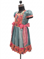 Happy young girl enjoying her premium blue and peach peacock motif silk knee-length ethnic frock inside a warm cream room - House of Kritee