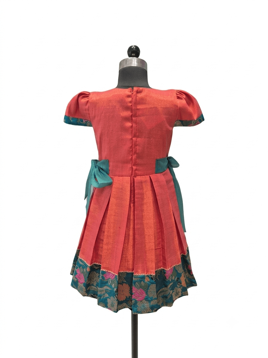 A short-sleeved dress displayed on a mannequin from the back, featuring a coral-red body, teal bow details at the waist, and a floral patterned border along the hem - House of Kritee