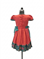 A short-sleeved dress displayed on a mannequin from the back, featuring a coral-red body, teal bow details at the waist, and a floral patterned border along the hem - House of Kritee