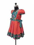 A traditional Indian salwar suit dress displayed on a black mannequin. The dress features a coral-pink cotton body with a teal floral brocade yoke, a matching teal waist tie, and a pleated skirt with a floral border.- House of Kritee