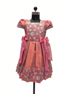A pink and coral printed dress displayed on a black mannequin against a white background. The dress features a floral pattern bodice, solid coral skirt panels, and large pink bow accents at the waist.- House of Kritee