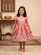 Happy young girl wearing a premium peach and pink floral brocade silk knee-length ethnic frock dress inside a warm cream room - House of Kritee