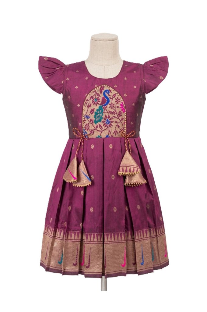 A purple traditional Indian girl's dress displayed on a mannequin. The dress features a round neckline, short flutter sleeves, a central peacock embroidery motif, and a pleated skirt with a decorative gold border and hanging tassels.
