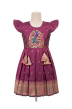 A purple traditional Indian girl's dress displayed on a mannequin. The dress features a round neckline, short flutter sleeves, a central peacock embroidery motif, and a pleated skirt with a decorative gold border and hanging tassels.