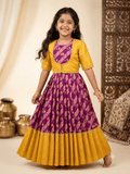 A young girl stands barefoot in a studio setting wearing a traditional Indian outfit consisting of a yellow kurta and a purple flared skirt with gold and pink patterns. The background features warm beige drapery, decorative brass pots, and a carved wooden screen.