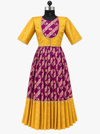 A young girl stands barefoot in a studio setting wearing a traditional Indian outfit consisting of a yellow kurta and a purple flared skirt with gold and pink patterns. The background features warm beige drapery, decorative brass pots, and a carved wooden screen.