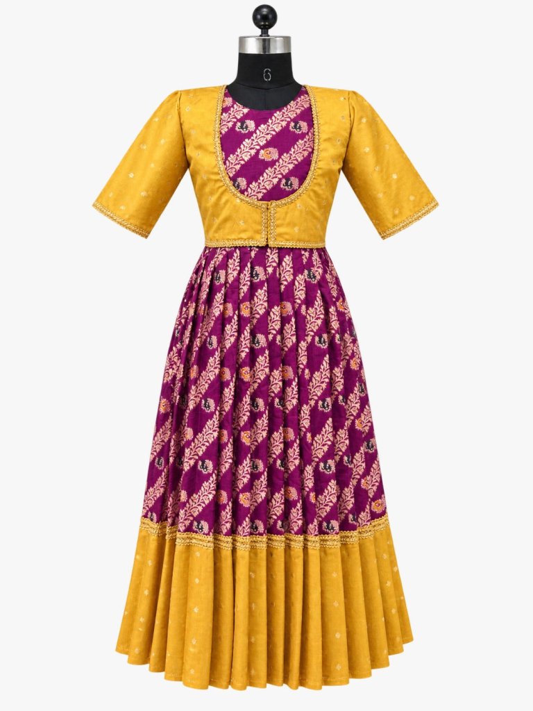 A young girl stands barefoot in a studio setting wearing a traditional Indian outfit consisting of a yellow kurta and a purple flared skirt with gold and pink patterns. The background features warm beige drapery, decorative brass pots, and a carved wooden screen.