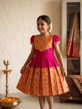 A young girl smiling and wearing a traditional Indian salwar kameez dress with a bright pink and orange patterned design, standing in an indoor setting with a brass lamp and a draped saree in the background. - House of Kritee
