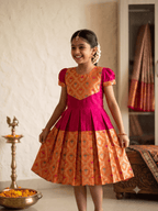 A young girl smiling and wearing a traditional Indian salwar kameez dress with a bright pink and orange patterned design, standing in an indoor setting with a brass lamp and a draped saree in the background. - House of Kritee