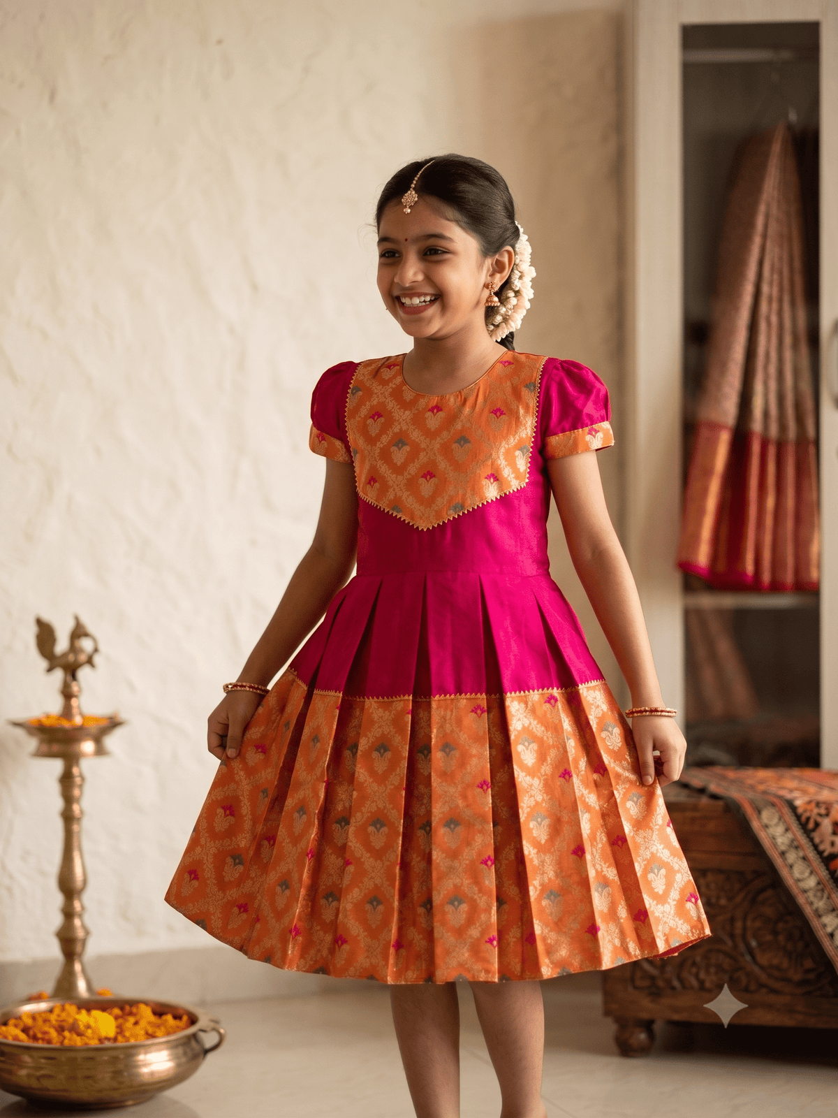 A young girl smiling and wearing a traditional Indian salwar kameez dress with a bright pink and orange patterned design, standing in an indoor setting with a brass lamp and a draped saree in the background. - House of Kritee