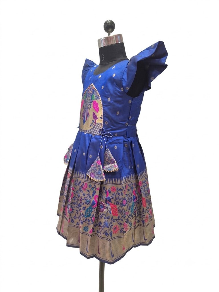 A royal blue sleeveless dress displayed on a black dress form mannequin against a white background. The dress features a heavily embroidered multicolor floral and geometric border on the skirt, a matching waist tie, and a central circular embroidered motif.