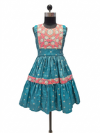 A sleeveless teal blue dress displayed on a black dress form mannequin, featuring a heavily embroidered coral pink and cream floral yoke and a tiered skirt with a matching coral pink band.