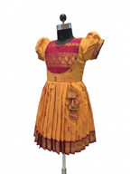 A traditional Indian girl's dress displayed on a black dress form mannequin. The dress features a mustard yellow body with a magenta-pink embroidered yoke, puffed sleeves, and a pleated skirt with a decorative tassel detail.