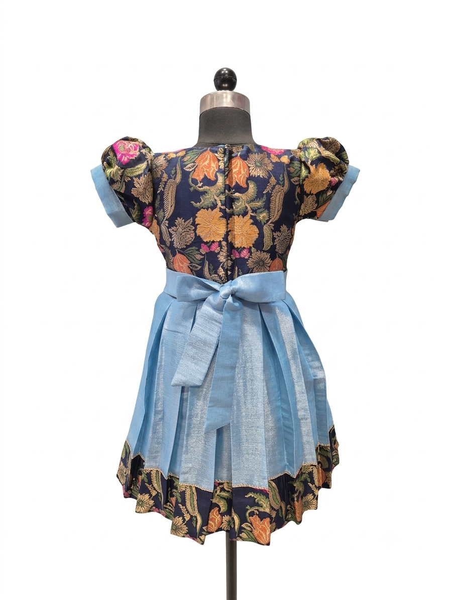 A short-sleeve dress displayed on a mannequin against a plain white background. The dress has a dark navy floral bodice with orange, yellow, and pink flowers, light blue puff sleeves, a matching light blue waist tie, and a light blue flared skirt with a dark floral ruffled hem.  - House of Kritee