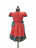 A short-sleeved dress displayed on a mannequin from the back, featuring a coral-red body, teal bow details at the waist, and a floral patterned border along the hem - House of Kritee