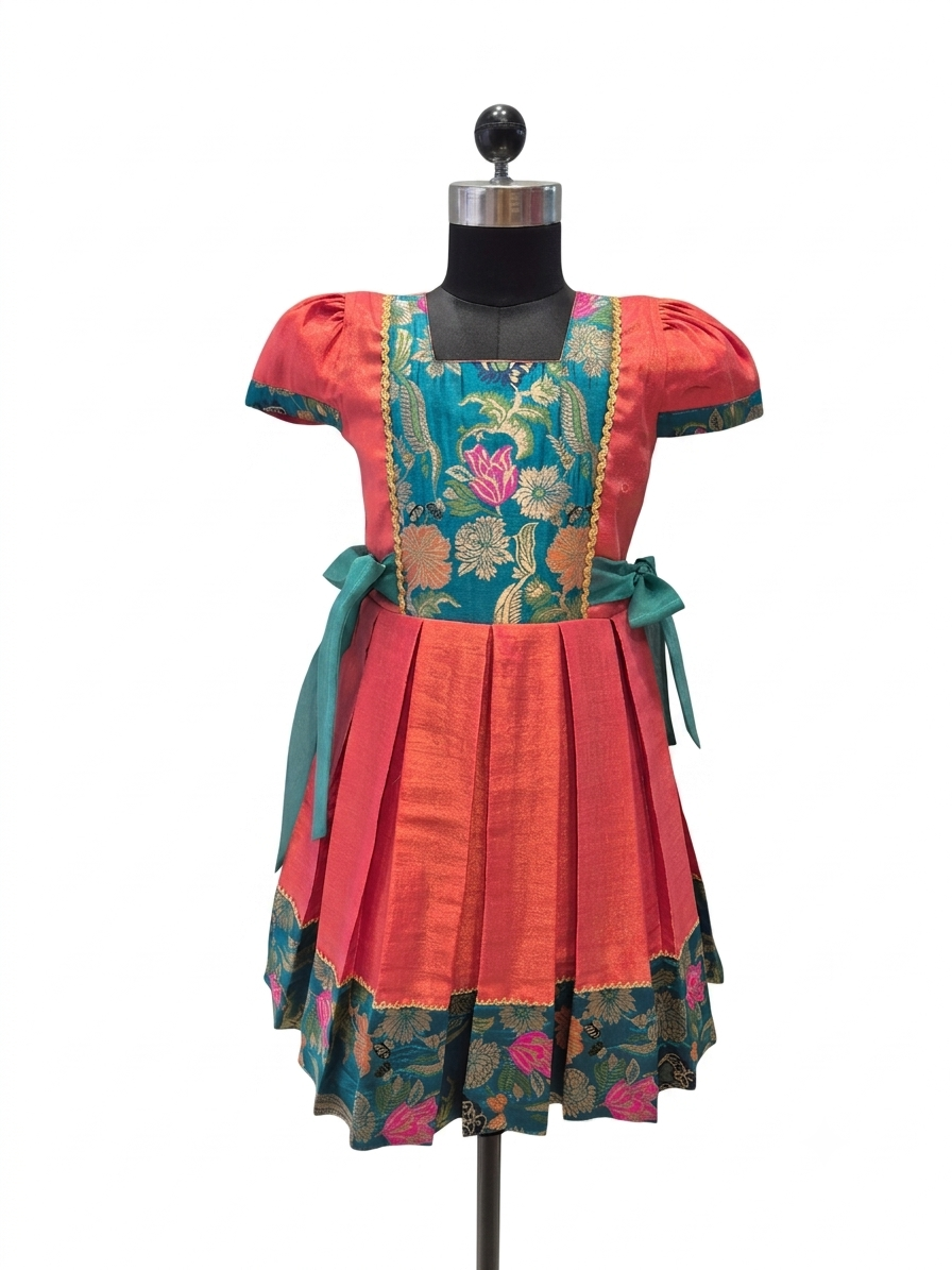 A short-sleeved traditional-style dress displayed on a black dress form against a plain white background. The dress has a coral-red skirt and sleeves with a teal floral brocade bodice and matching teal ribbon ties at the waist.- House of Kritee