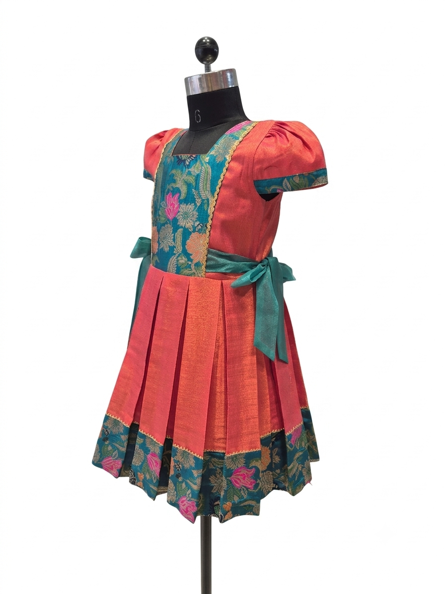 A traditional Indian salwar suit dress displayed on a black mannequin. The dress features a coral-pink cotton body with a teal floral brocade yoke, a matching teal waist tie, and a pleated skirt with a floral border.- House of Kritee