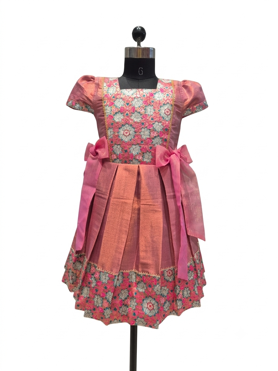 A pink and coral printed dress displayed on a black mannequin against a white background. The dress features a floral pattern bodice, solid coral skirt panels, and large pink bow accents at the waist.- House of Kritee