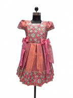 A pink and coral printed dress displayed on a black mannequin against a white background. The dress features a floral pattern bodice, solid coral skirt panels, and large pink bow accents at the waist.- House of Kritee