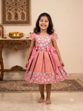 Happy young girl wearing a premium peach and pink floral brocade silk knee-length ethnic frock dress inside a warm cream room - House of Kritee