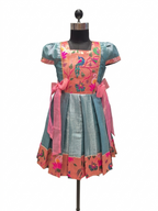 A short-sleeved traditional-style dress displayed on a black dress form mannequin against a plain white background. The dress features a teal blue lower skirt, a peach-pink upper bodice with embroidered peacock and floral motifs, and pink ribbon ties at the waist. - House of Kritee